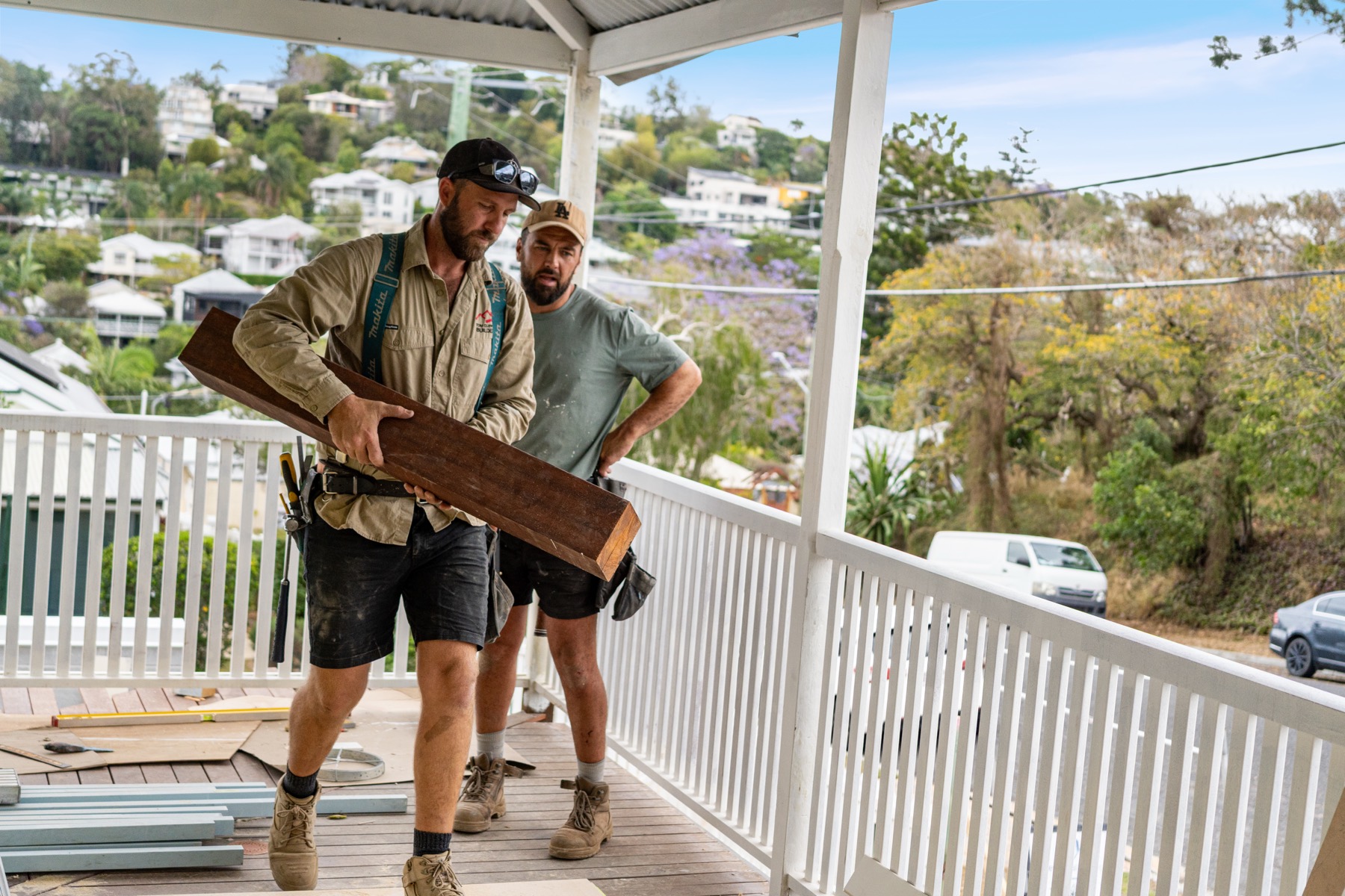 Two carpenters carrying a length of hardwood across a deck above Brisbane's hills